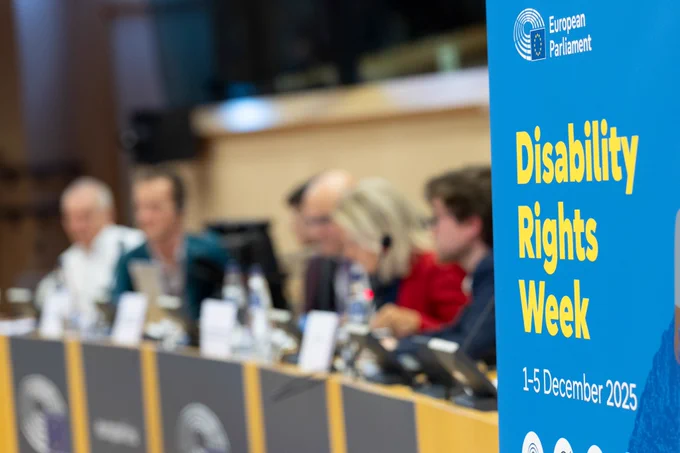 Close up of European Parliament's Disability Rights Week 2025 roll up. In the blurry background, 6 speakers of the Transport Committee event mobility for persons with disabilities.
