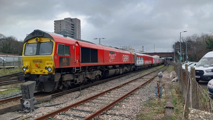 A red GBRf freight train with white and blue carriages parked on a railway track, with cars parked alongside and buildings in the background.