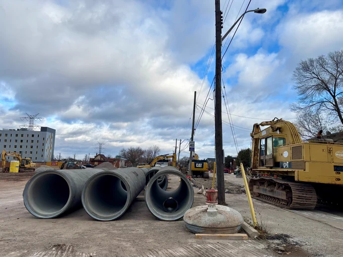 Un chantier de construction avec de grands tuyaux en béton au sol, une excavatrice Caterpillar jaune à droite et un ciel partiellement nuageux au-dessus.
