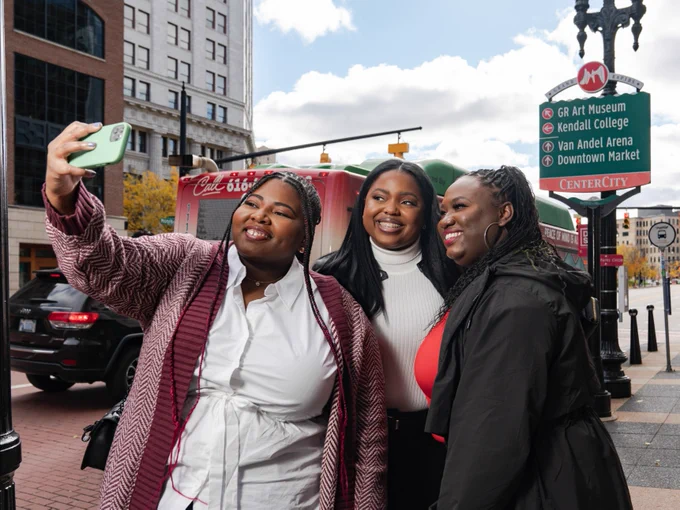 Three individuals taking a selfie together on a city street, with a directional sign in the background pointing to various landmarks in Grand Rapids.