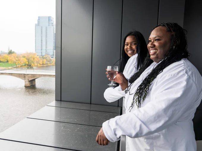 Two individuals in white outfits smiling and holding wine glasses on a balcony, with a cityscape and a river in the background.