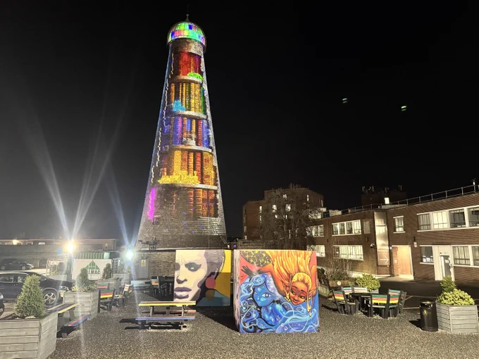 St. Patrick's Tower is illuminated with colorful lights standing tall at night on The Digital Hub's campus. In front, vibrant graffiti artwork cubes feature faces and abstract designs. Colourful benches are evident on the gravel beside the cubes.