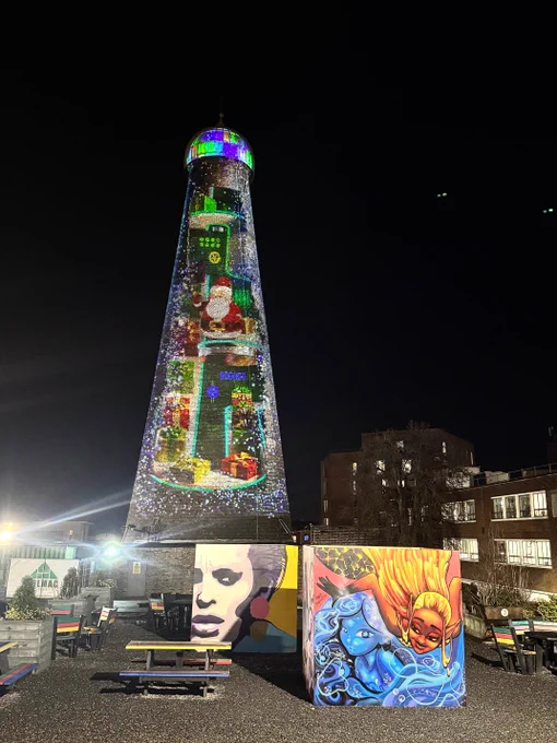 St. Patrick's Tower is illuminated with colorful lights standing tall at night on The Digital Hub's campus. In front, vibrant graffiti artwork cubes feature faces and abstract designs. Colourful benches are evident on the gravel beside the cubes.