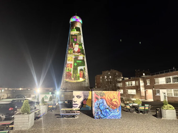 St. Patrick's Tower is illuminated with colorful lights standing tall at night on The Digital Hub's campus. In front, vibrant graffiti artwork cubes feature faces and abstract designs. Colourful benches are evident on the gravel beside the cubes.