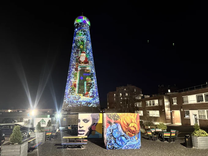St. Patrick's Tower is illuminated with colorful lights standing tall at night on The Digital Hub's campus. In front, vibrant graffiti artwork cubes feature faces and abstract designs. Colourful benches are evident on the gravel beside the cubes.