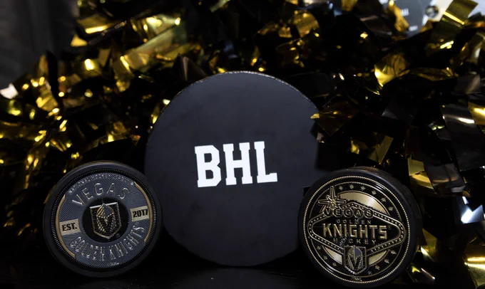 The BHL hockey puck is in the middle between two Vegas Golden Knight, hockey pucks and there is gold streamers behind as decoration