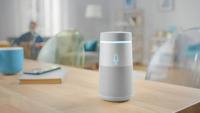 A cylindrical smart speaker sits on a wooden table in a living room. 