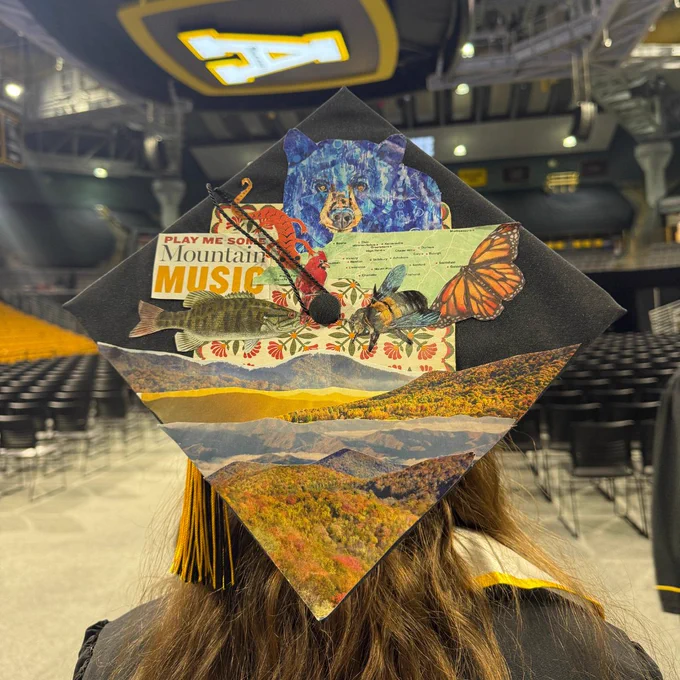 Graduate wearing a decorated cap featuring a collage of mountains, a map, a butterfly, a cardinal, a bear, a fish and a bee, alongside a quote that reads “Play me some Mountain Music”. Seating for a graduation ceremony can be seen in the background. 