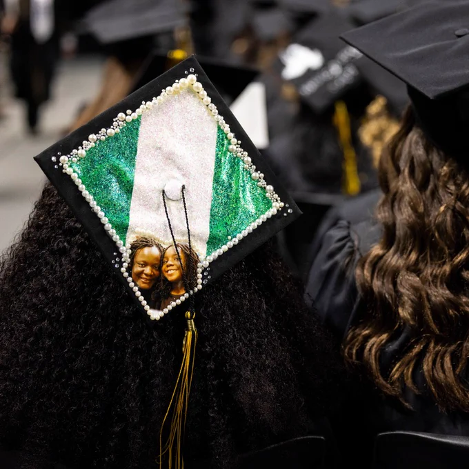 Graduate wearing a decorated cap with a photograph and the Nigerian flag at a graduation ceremony.