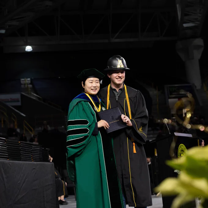 A graduate on stage receiving their diploma from a faculty member. The graduate is wearing a black hard hat with an App State A on the front. The faculty member is wearing dark green regalia. Both are smiling for a photo. 