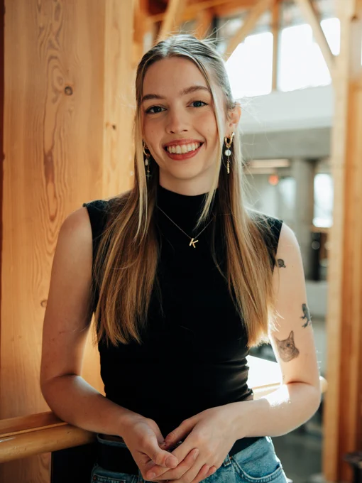 Person smiling in a sunlit room, leaning against a wooden railing, wearing a black top and denim jeans, accessorized with earrings and a necklace.
