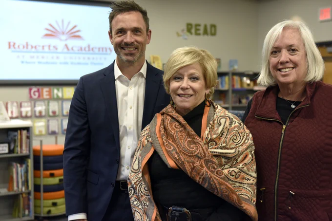 Three individuals smiling in a library with a sign for Roberts Academy in the background.