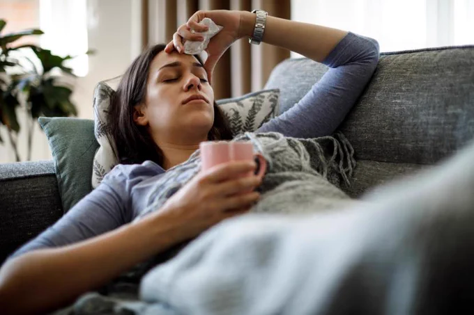 Person resting on a couch, holding a mug and a tissue, with eyes closed.