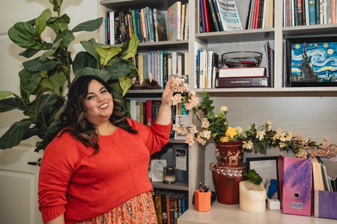 Jennifer van Alstyne, a latina woman, stands in her home office in San Diego by a built-in bookcase filled with books. She's lightly touching the tip of a branch of cherry blossom flowers leaning out of a llama vase.