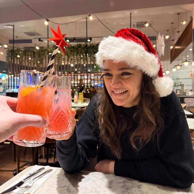 Person wearing a Santa hat smiling and toasting with a drink at a restaurant table.