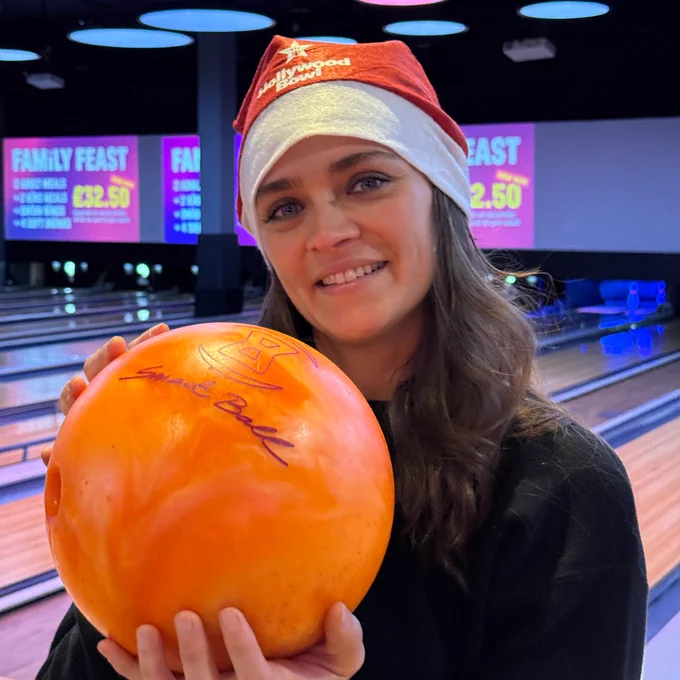 Person wearing a Hollywood Bowl Santa a hat holds a signed orange bowling ball at a bowling alley.