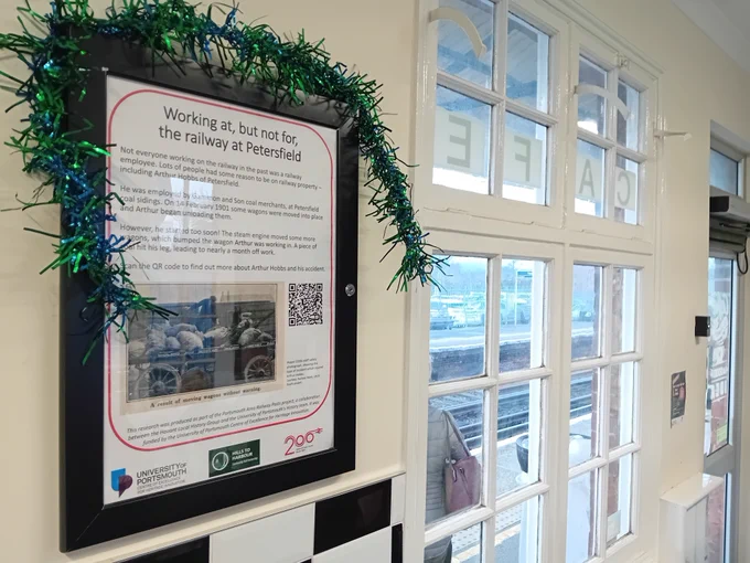 Photograph of a poster in a frame, titled 'Working at, but not for, the railway at Petersfield', on a waiting room wall, with a window beyond. Through the window platform and tracks can be seen. A piece of green tinsel sits over the poster frame.
