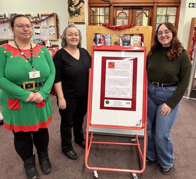 Three women stand indoors beside a red-framed display board featuring a framed piece of writing. The setting appears to be a library, with bookshelves, noticeboards, and Christmas decorations in the background. One woman wears a green elf-themed dress, another is dressed in black, and the third wears a dark green top and jeans. All three are smiling and facing the camera.