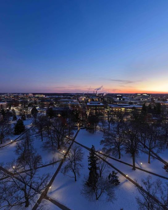 Aerial shot of a snow-covered MSU campus at dusk with bare trees lining intersecting paths. Buildings and city lights are in the background. The sky transitions from blue to orange on the horizon.