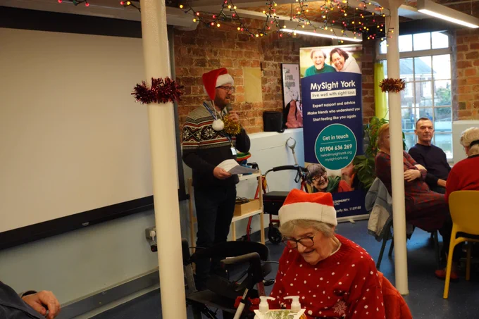 Scott, MySight York's Chief Executive is wearing a Santa hat, addressing the room whilst holding a mic.