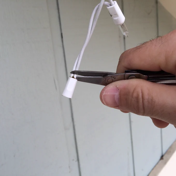 Small scissors cutting the white plastic socket from a string light cord while the bulb hangs above.