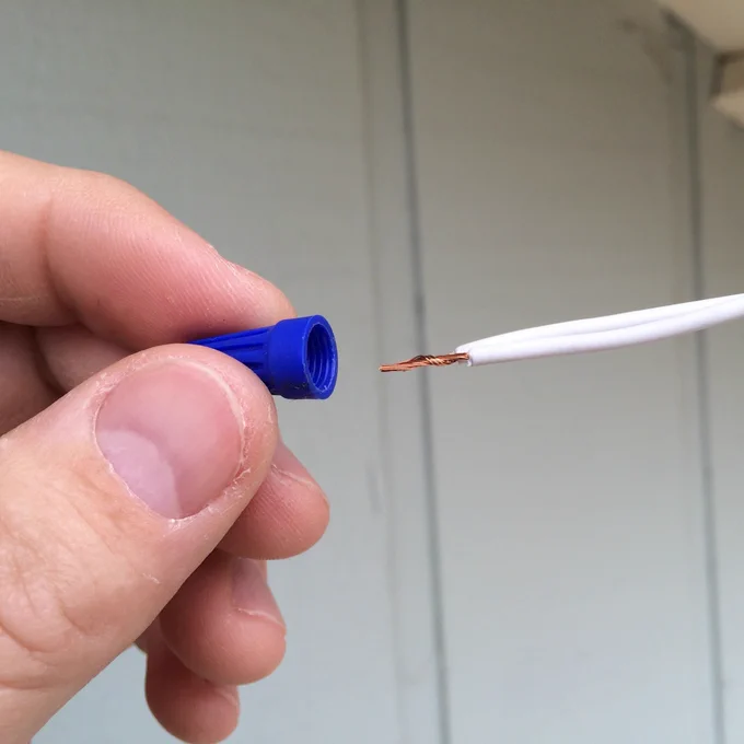 Close-up of a hand holding a blue wire nut next to a white electrical wire with exposed copper strands.