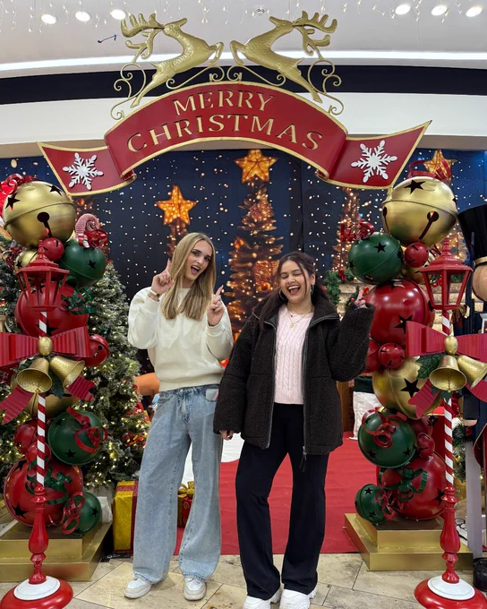 Two people standing and smiling in front of a festive "Merry Christmas" display, featuring a large ornamental sign, Christmas trees, and decorative reindeer and nutcracker statues