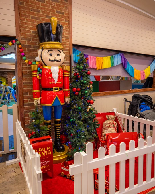 Large nutcracker statue standing next to a decorated Christmas tree inside a room, with a sign reading 'Santa's Workshop North Pole' and colourful streamers in the background.