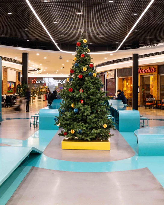 Decorated Christmas tree centered in a bustling shopping mall with seating areas and storefronts, including Nando's, visible in the background.