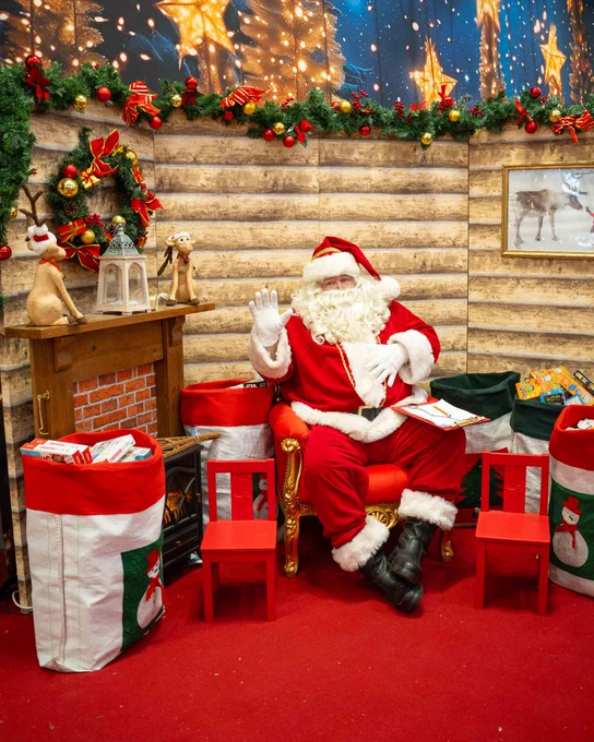 Santa Claus seated in a large chair, waving in a festively decorated Christmas setting with garlands, lights, and presents around, capturing the holiday spirit.
