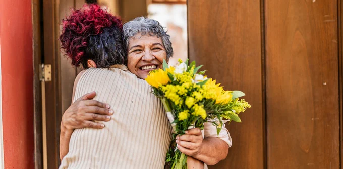 Two individuals embracing joyfully; one holds a bouquet of yellow flowers, both are smiling. They stand in front of a wooden door.