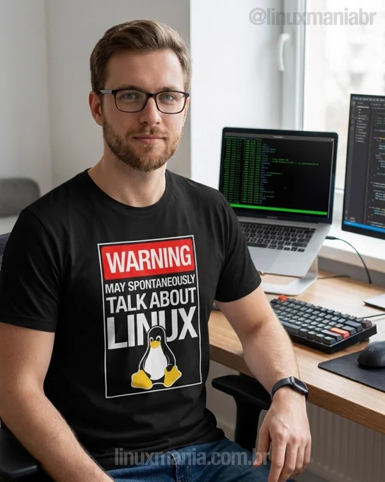 Homem branco, de barba e óculos, sentado em uma mesa de trabalho com notebook exibindo código, usando camiseta preta com a estampa “WARNING: May spontaneously talk about Linux” e o mascote Tux.