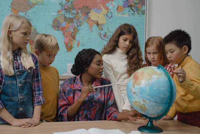 Teacher points to a globe to a group of attentive children in a classroom with a world map behind them.