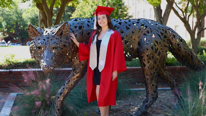 Josie stands in front of a wolf statue in her graduation regalia.
