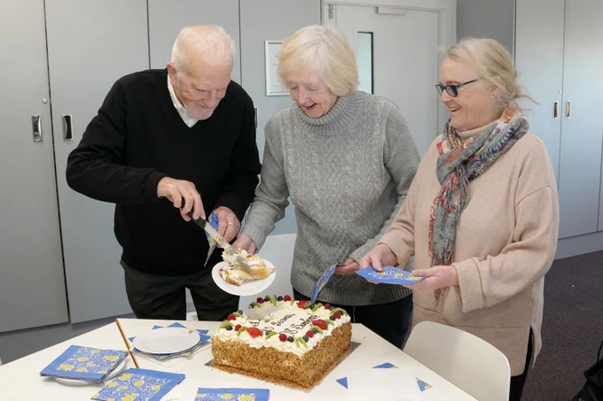 Three people joyfully cut and serve a large decorated cake in a bright room, celebrating a special occasion together. Paper plates and napkins are placed on the table for serving.