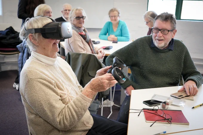 A person wearing a VR headset holds a controller, smiling, while another person sitting at the table watches, surrounded by other people sitting at tables in a classroom.