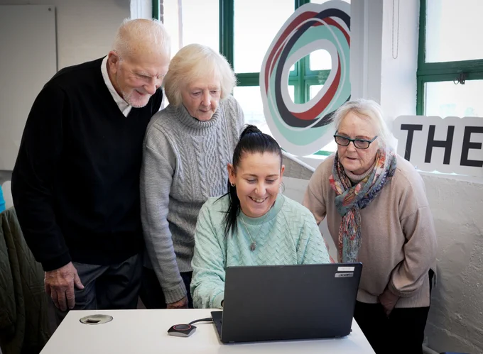 A smiling person helps three people learn to use a laptop in a bright room, fostering a sense of community and learning.