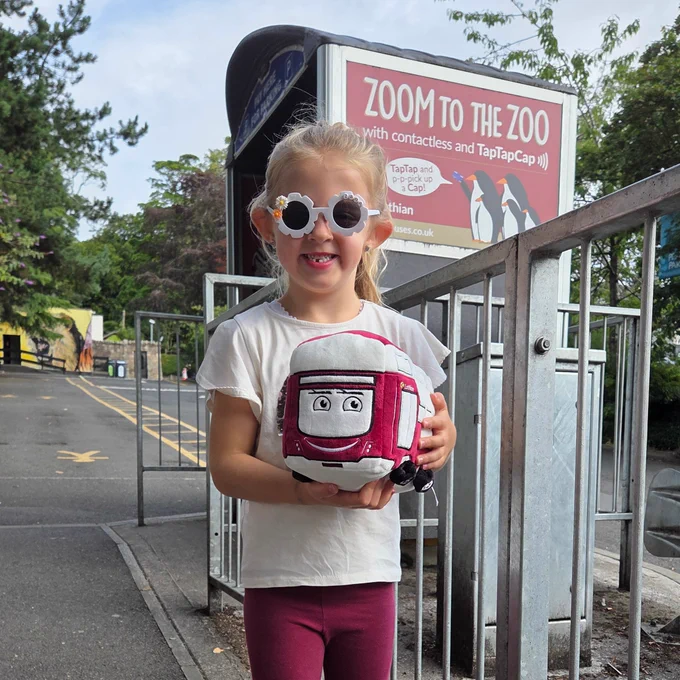 Child holding a plush toy of a red and white bus standing near a metal railing outside on a paved path.