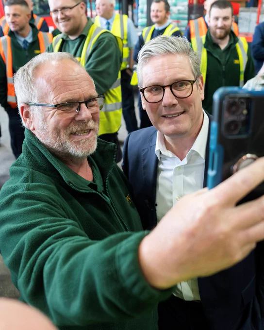 Prime Minister Keir Starmer stands next to a worker taking a selfie. Other workers in PPE stand behind them.