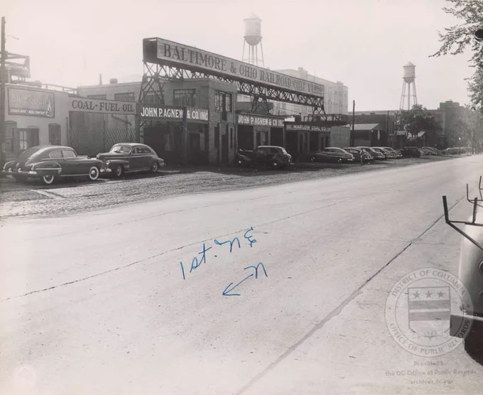 A black and white image of the Baltimore and Ohio Railroad Coal Yard on 1st Street NE in Washington, DC. There are several cars parked in front of the building and two water towers can be seen in the distance. 