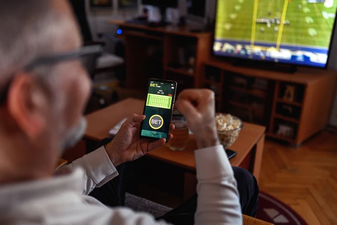 Older gentleman sitting on the sofa at home and watching a football game. He is holding a smart phone and using it for sports betting.