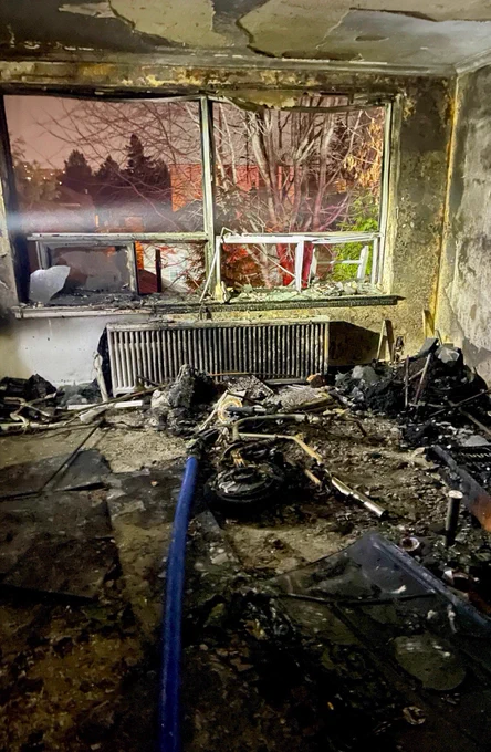 Image of a fire damaged room. A blue fire hose sitting on the floor in the middle of the room. The remnants of an e-bike in the middle of the room at the end of the hose nozzle.