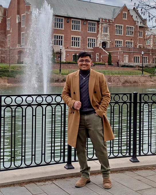 A person stands smiling in front of a fountain and a traditional brick building, dressed in a long coat and glasses.