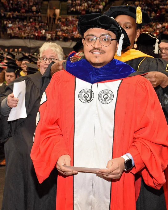 Graduate in red and black academic regalia holding a diploma at a commencement ceremony.