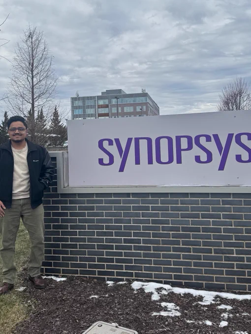 Person standing beside a Synopsys sign outside, with a building and trees in the background.