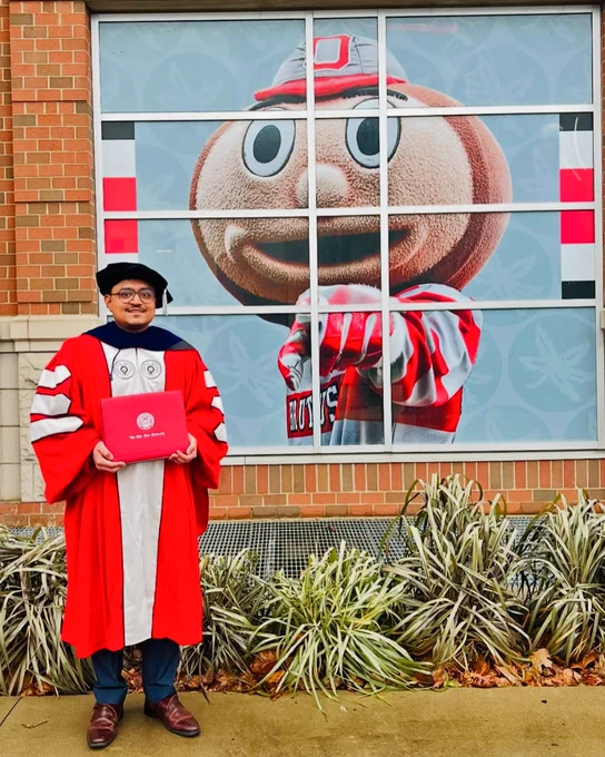A graduate in red and white academic regalia holds a diploma in front of a large window display featuring Ohio State University's mascot, Brutus Buckeye.