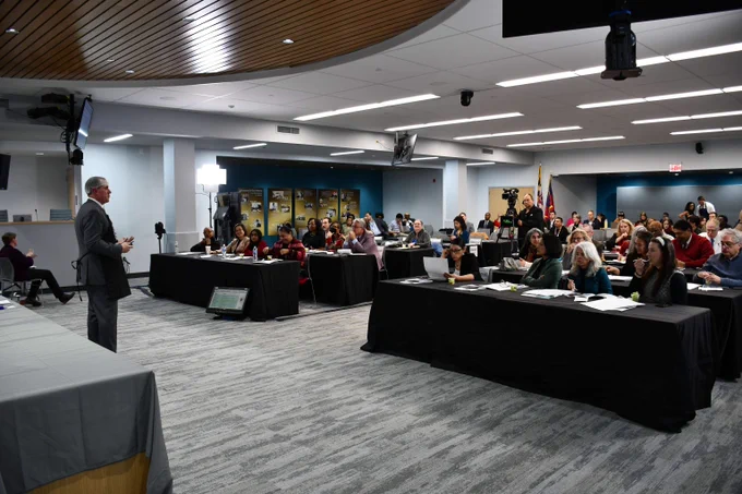 Speaker addressing a large audience seated at tables in a modern conference room with ceiling lights and wood paneling.