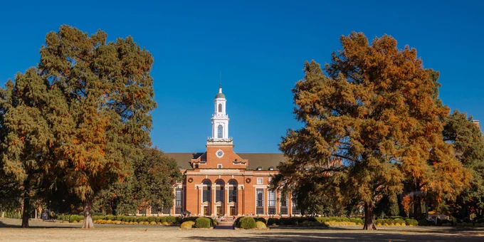 View of Edmon Low Library at Oklahoma State University, featuring a classic red-brick building with a central white clock tower, surrounded by large trees with autumn foliage under a clear blue sky.