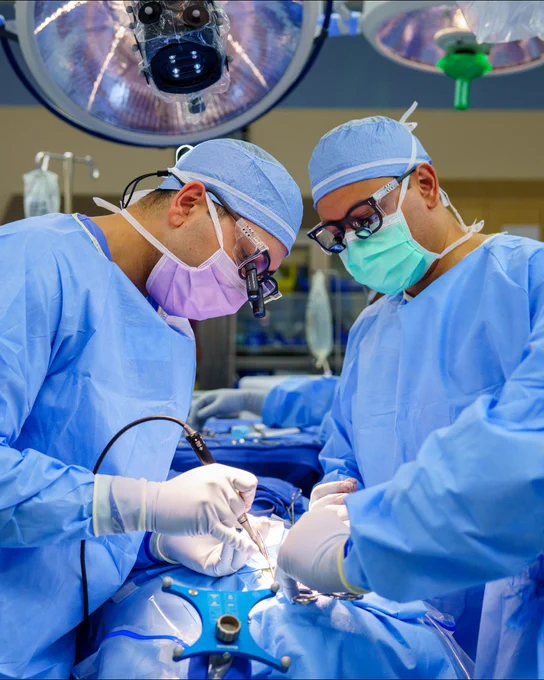 Two surgeons in scrubs and surgical masks are focused on a procedure in an operating room, with bright overhead lights illuminating the surgical area.