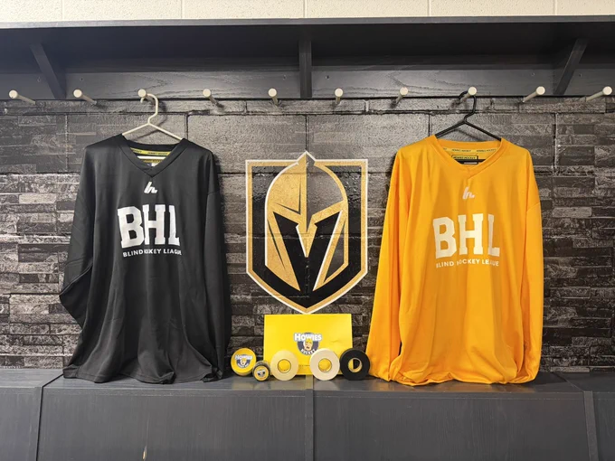 A black jersey and a yellow jersey both with a BHL logo hang in the locker room stall inside city national Arena with the Vegas Golden Knights logo in the middle. Various types of howies hockey tape is placed in the middle.